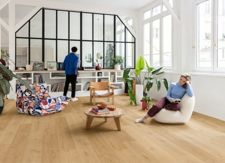 lvt-floors-quick-step-bloom-elegant-oak-natural LVT floors Quick-Step Bloom Elegant Oak Natural installed on a living room floor. Woman is sitting on a chair and reading a book. Man is looking the vinyl record player. Table with books, palm threes