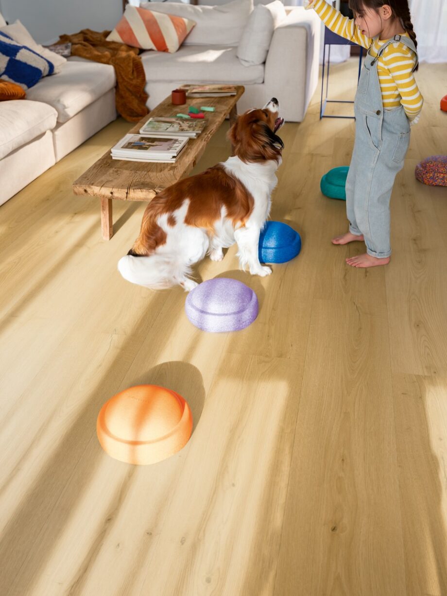 vinyl-quick-step-bloom-brushed-oak-honey Vinylboden Quick Step Bloom Brushed Oak Honey im Wohnzimmer verlegt, Kind spielt mit einem Hund.