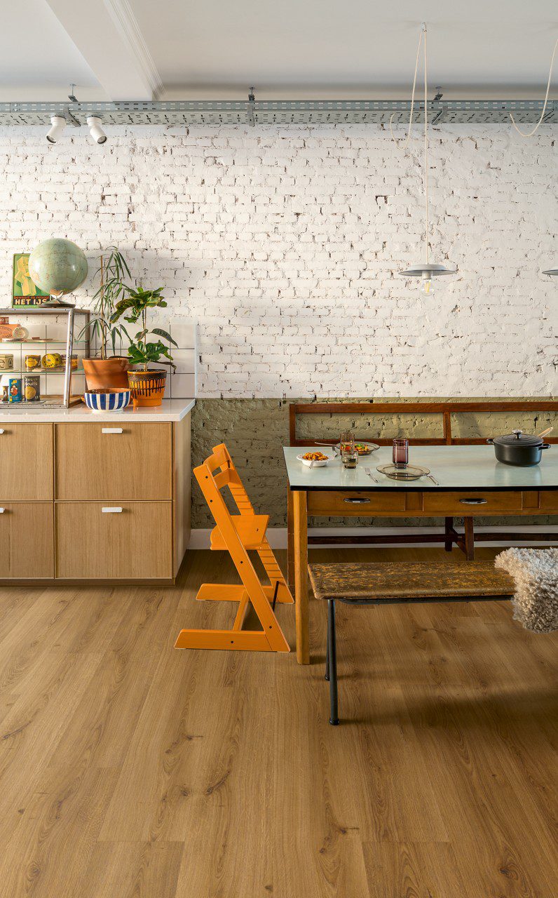 Vinylboden Quick Step Bloom Botanic Caramel Oak im Esszimmer verlegt, orangefarbener Kinderstuhl, Esstisch, Schränke und Blumen.