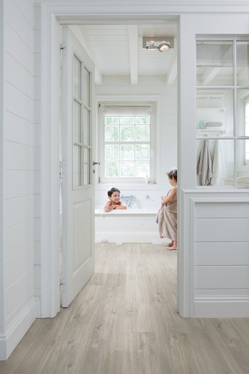 Vinyylilattia Quick Step Blos Vaalea Canyon Tammi Saw Cuts asennettuna makuuhuoneen ja kylpyhuoneen lattialle, lasiovi avoinna kylpyhuoneeseen, vaahtokylpy jossa yksi lapsi leikkii ja toinen seisoo lattialla vaahtoisin jaloin.