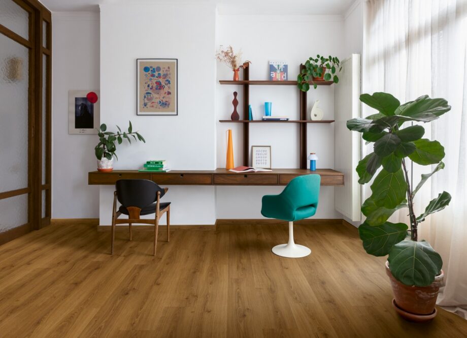 виниловыи-ламинат-quick-step-bloom-botanic-caramel-oak Виниловый ламинат Quick Step Bloom Botanic Caramel Oak уложен на полу в домашнем офисе, письменный стол, светло-голубое офисное кресло, пальма и цветы.