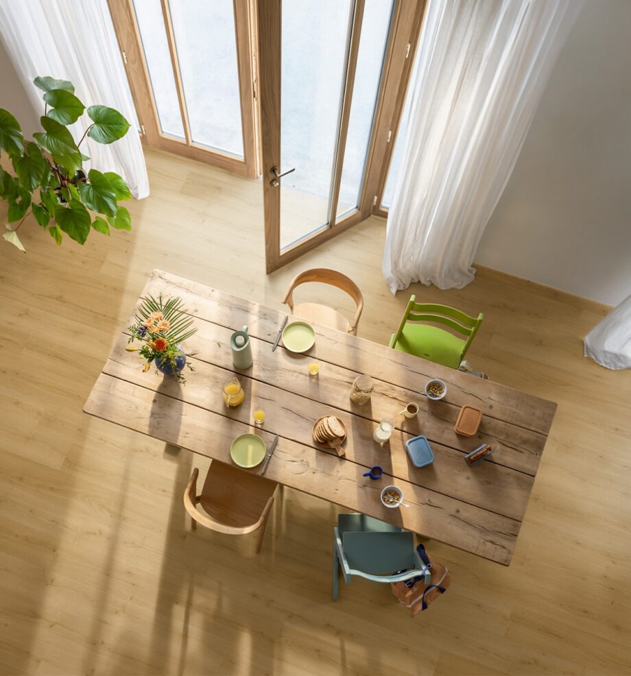 виниловый-паркет-quick-step-bloom-brushed-oak-honey Виниловый паркет Quick Step Bloom Brushed Oak Honey уложен на полу на кухне, обеденный стол со стульями и открытая дверь на террасу.