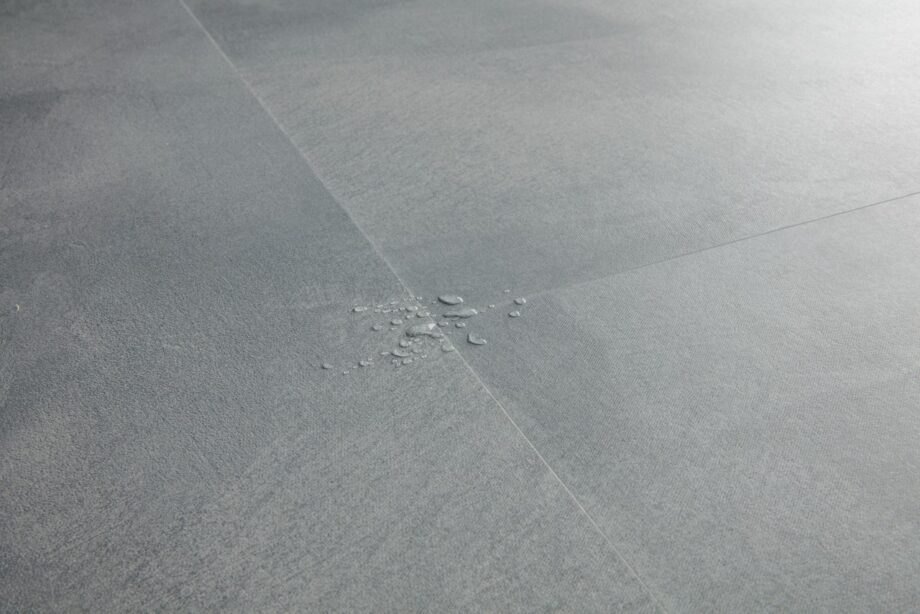 Vinyylilaatta Quick-Step Illume Soft Sky asennettuna kodin lattialle – vaalea, raikas harmaansininen sävy ja hillitty kivimäinen/betonimainen design hienovaraisella tekstuurilla; vinüylipinnan vesipisarat korostavat lattian vedenkestävyyttä.