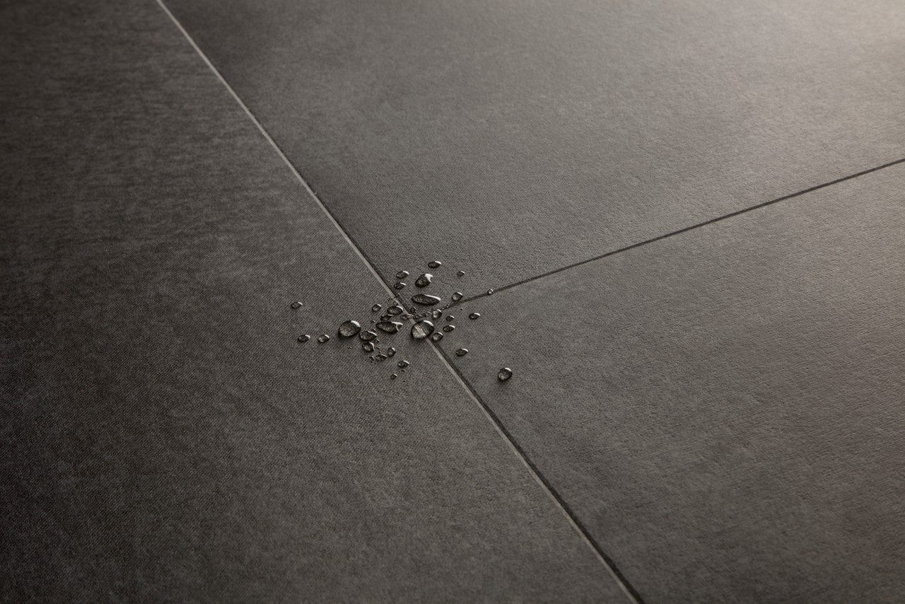 Vinyylilattia Quick-Step Illume Soft Graphite asennettuna kodin lattialle – tumma grafiitinharmaa sävy ja hillitty kivimäinen/betonimainen design hienovaraisella tekstuurilla; lattian pinnalla näkyy vesipisaroita korostamassa sen vedenkestävyyttä.