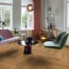 Helles, stilvolles Wohnzimmer mit Fischgrätparkett aus Eiche Mix (140 mm, geölt). Die Einrichtung kombiniert zartrosa Sofa, salbeigrünen Designstuhl und einen modernen Couchtisch mit goldenen Akzenten. Große weiße Flügeltüren und Fenster bringen viel Tageslicht in den Raum. Warme Vorhänge, moderne Kunstobjekte und eine elegante Ambienteleuchte schaffen eine harmonische und einladende Atmosphäre. Herringbone flooring Oak Mix oil-waxed 140 mm – 9