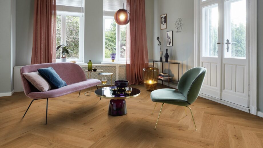Helles, stilvolles Wohnzimmer mit Fischgrätparkett aus Eiche Mix (140 mm, geölt). Die Einrichtung kombiniert zartrosa Sofa, salbeigrünen Designstuhl und einen modernen Couchtisch mit goldenen Akzenten. Große weiße Flügeltüren und Fenster bringen viel Tageslicht in den Raum. Warme Vorhänge, moderne Kunstobjekte und eine elegante Ambienteleuchte schaffen eine harmonische und einladende Atmosphäre. Herringbone flooring Oak Mix oil-waxed 140 mm – 9