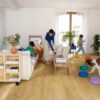 lvt-parkett-quick-step-bloom-brushed-oak-honey LVT parkett Quick-Step Bloom brushed oak honey paigaldatud elutoa põrandale. Ema koos kahe lapsega, koer, diican, lilled, riiul, telekalaud.