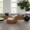 lvt-parkett-quick-step-bloom-cotton-oak-cozy-grey LVT parkett Quick-Step Bloom Cotton Oak Cozy Grey paigaldatud elutoa põrandale. Pruunist nahast dugitool, diivanilaud lilledega, palmipuu, pikk kummut akende all.