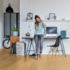 LVT Quick Step Bloom Pure Oak Honey paigaldatud kodukontori põrandale. Naine räägib telefoniga toetudes kontori lauale. Tool, jalgratas, apple arvuti.