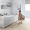 Veekindel vinüülparkett Quick-Step Blos Canyon tamm beige paigaldatud vannitoa põrandale. Valge kandiline vann on täis vannivahtu ja väike tüdruk on vannist välja tulnud ning on käterätis. Põrand on koos vannivahuse veega. Kuivaturedel millel on särk kuivamas ja seinal on pilt liivasest rannast koos kasvavate heintega.