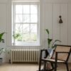 vinly-floor-quick-step-blos-buttermilk-oak Quick Step Blos Buttermilk Oak vinyl floor installed in a living room, with a classic wall radiator, a rocking chair, white classic wall panels, and potted plants.