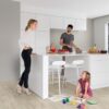 Vinüülparkett Quick-Step Blos Canyon tamm beige paigaldatud köögi põrandale. Valge köögimööbel ja köögisaar millel isa lõikab perele salatit. V'ike tütar joonistab õrandal värvidega pilte ja ema jälgib teda toetudes käega köögisaarele.