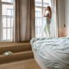 Vinyylilattia Quick Step Bloom Elegant Fumed Oak asennettuna makuuhuoneen lattialle, tumma savutammen sävy ja lankkudesign, suuri sänky ja nainen juo aamukahvia katsoen ikkunasta ulos.