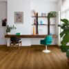 виниловыи-ламинат-quick-step-bloom-botanic-caramel-oak Виниловый ламинат Quick Step Bloom Botanic Caramel Oak уложен на полу в домашнем офисе, письменный стол, светло-голубое офисное кресло, пальма и цветы.