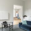 Vinylboden in Betonoptik Quick-Step Illume Soft Sky, verlegt auf dem Wohnzimmerboden – schwarzer Ledersessel und blaues Sofa; im Hintergrund sind in der Küche ein stehender Vater und ein kleiner Sohn auf einem Dreirad zu sehen.