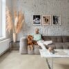 vinylboden-betonoptik-quick-step-illume-sandstone-concrete Vinylboden in Betonoptik, verlegt auf dem Wohnzimmerboden – weiß gestrichene rustikale Ziegelwand, Couchtisch aus weißem Marmor mit verchromten Metallbeinen und graues Sofa; eine blonde Frau streichelt ihren braunen Hund.